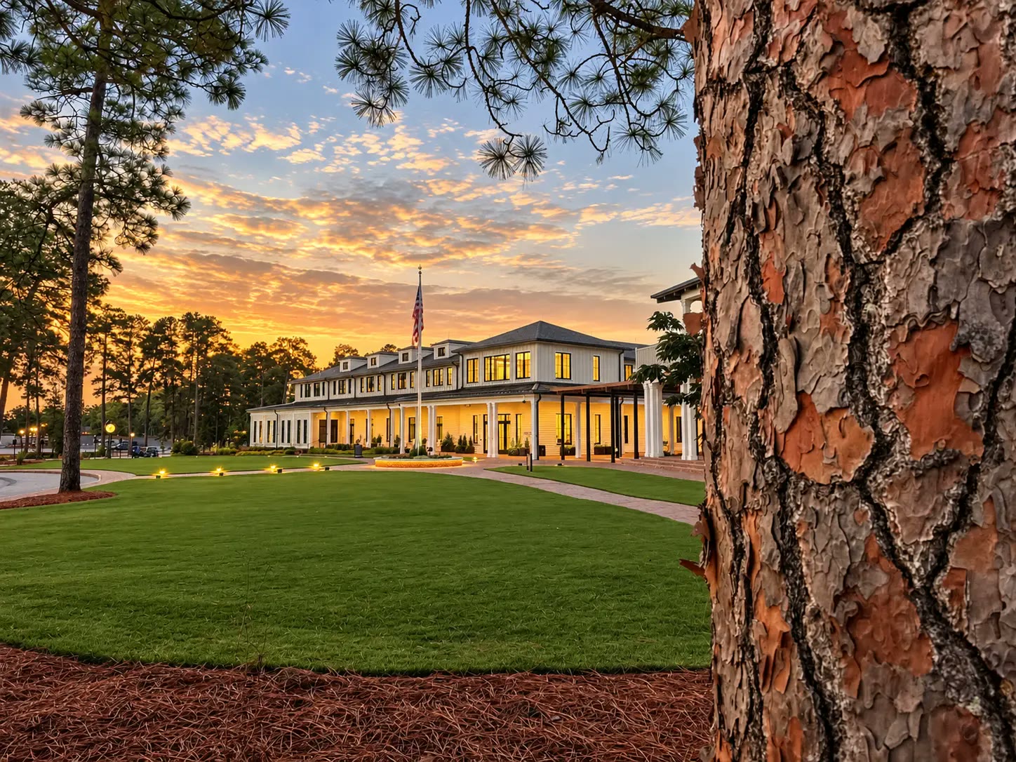 Golf House Pinehurst building lit at twilight beneath longleaf pines, with an American flag and lawn in front